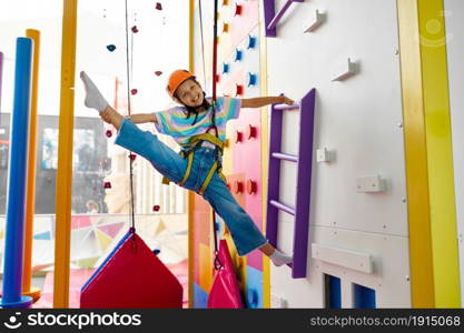 Cute girl in helmet is hanging on climbing wall, entertainment center, young climber. Children having fun, kids spend the weekend on playground, happy childhood. Cute girl in helmet is hanging on climbing wall