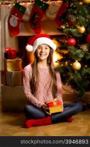 Cute girl in Santa cap sitting on floor with Christmas present at fireplace