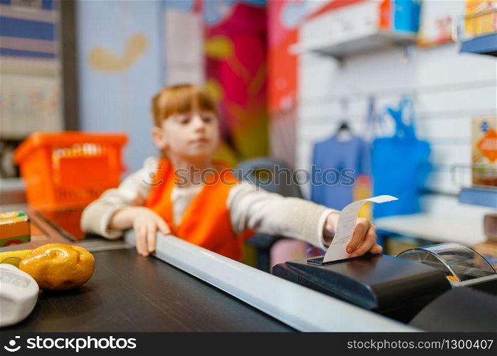 Cute girl in uniform at the cash register makes a check for the purchase, saleswoman, playroom. Kids plays sellers in imaginary supermarket, salesman profession learning. Cute girl at the register makes check for purchase