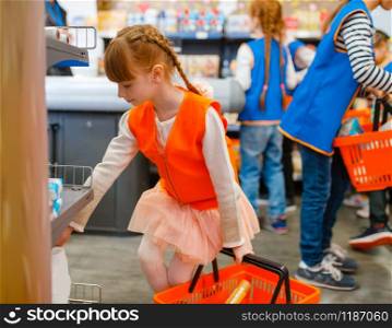 Cute girl in uniform with basket playing saleswoman, playroom. Kids plays sellers in imaginary supermarket. Cute girl with basket playing saleswoman, playroom