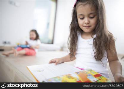 Cute girl reading book with sister in background at home