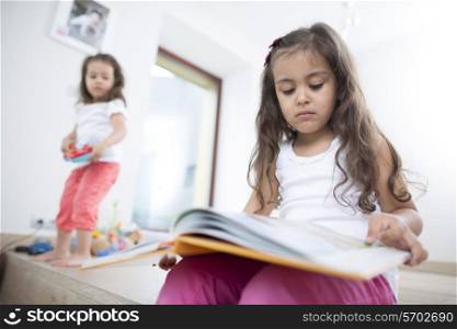 Cute girl reading book with sister playing in background at home