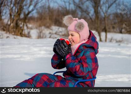 Cute girl with a red cup sits in a sleigh in the snow. The child warms himself with hot tea on a sunny winter day.. Cute girl with a red cup sits in a sleigh in the snow. The child warms himself with hot tea.