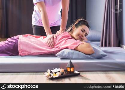 Cute girl with traditional dress massaging back by professional with candle and Plumeria on bed. portrait of happy Asian beautiful woman relax in spa salon. Body care treatment by Thai massage.