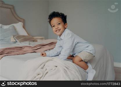 Cute happy african american kid boy playing and having fun on bed after coming home from kindergarten, playing and smiling while spending time in bedroom. Carefree childhood concept. Cute young afro american boy playing on big bed at home