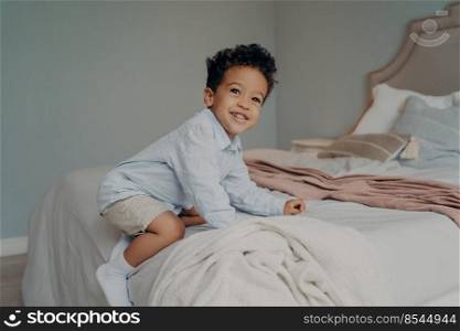 Cute happy african american kid boy playing and having fun on bed after coming home from kindergarten, playing and smiling while spending time in bedroom. Carefree childhood concept. Cute young afro american boy playing on big bed at home