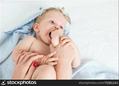Cute happy beautiful infant boy playing with his foot with wet hair sitting in hothouse bath light blue fluffy towel white background, horizontal photo.