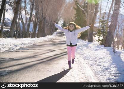 Cute happy little girl having fun in winter park, running along road with raised up hands, enjoying bright sunny day in wintertime