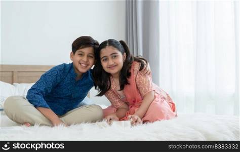 Cute Indian asian siblings with traditional clothing sitting on bed, handsome brother embracing beautiful younger sister, smiling and looking at camera while playing together at home. Family lifestyle