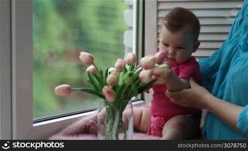 Cute infant child sitting on window sill and playing with tulip flowers. Adorable baby girl trying to bite tulip. Young mother holding little kid and showing flowers to the child while relaxing togehter at home.
