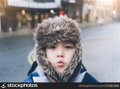 Cute Kid boy wearing a fur hooded winter coat standing alone with blurry light bokeh and glowing sunset shining, Emotional portrait child feeling cold while walking on street in the city in evening