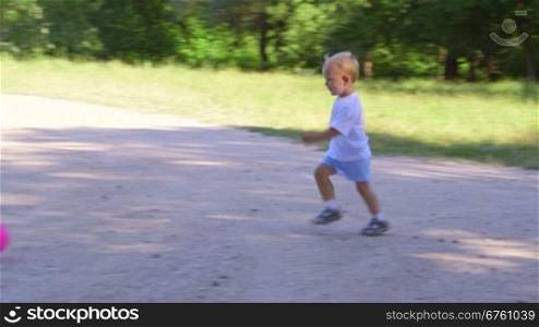 Cute kid runs and kicks the ball on the footpath in summer park