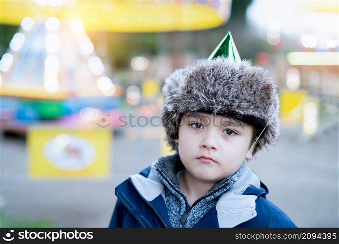 Cute kid with fluffy party hat looking out deep in thought, Young boy standing alone outside with blurry colourful light in playground background, Child at School Christmas party on Winter holiday