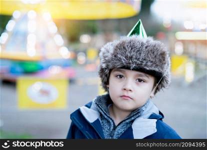 Cute kid with fluffy party hat looking out deep in thought, Young boy standing alone outside with blurry colourful light in playground background, Child at School Christmas party on Winter holiday