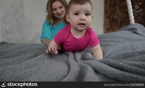 Cute laughing infant child learning to crawl on bed. Caring mother playing with her baby girl in bedroom while spending leisure at home. Adorable newborn kid crawling on bed and smiling. Slow motion.