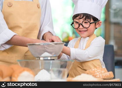 Cute little Asian boy and beautiful mother sifting dough flour with sifter sieve colander in home kitchen on table for prepare to baking bakery and cake. Thai kids playing with flour as chef funny