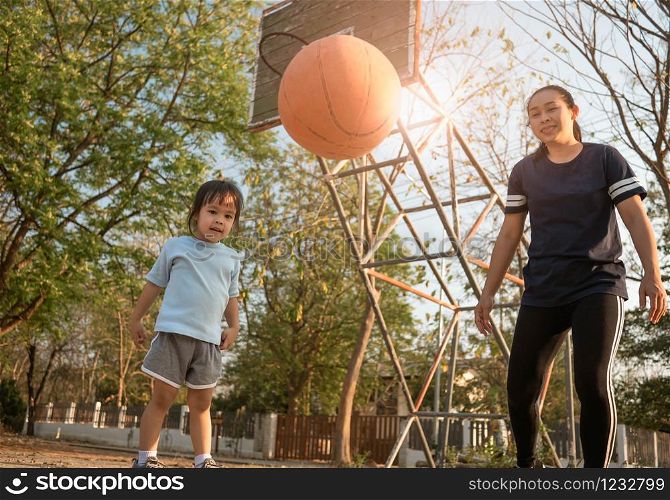 Cute little Asian child playing basketball with her mother on playground on summer day. Happy family spending free time together.