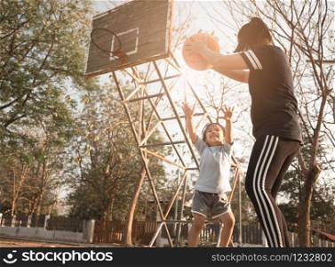 Cute little Asian child playing basketball with her mother on playground on summer day. Happy family spending free time together.
