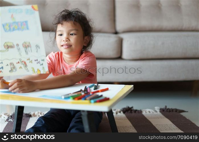 Cute little asian girl Sitting holding a drawing paper, drawing homework and writing with color Wax crayons on paper at living room
