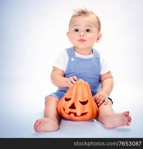 Cute little baby boy sitting in the studio over clear background with carved orange pumpkin, happy Halloween day, traditional American autumn holiday