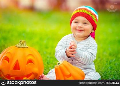 Cute little baby boy wearing funny colorful hat, playing with two beautiful carved pumpkins, traditional Halloween symbol, having fun on sunny autumnal day