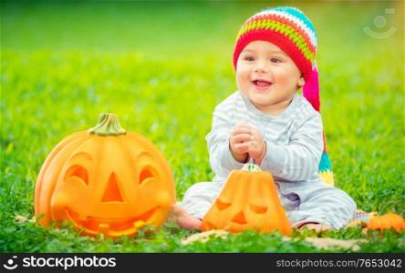 Cute little baby boy wearing funny colorful hat sitting in backyard on green grass meadow near two pumpkins, playing outdoors with traditional Halloween toys