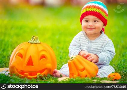 Cute little baby boy wearing funny colorful hat, sitting in backyard on green grass meadow near two decorative pumpkins, playing outdoors with traditional Halloween toy