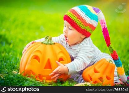 Cute little baby boy wearing funny colorful hat trying to bite beautiful carved festive pumpkin, playing outdoors with traditional Halloween toys