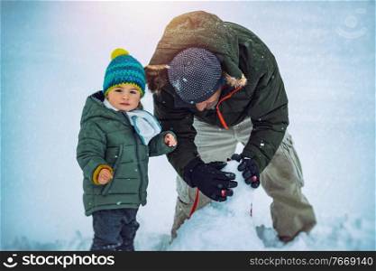 Cute Little Baby Boy with Father Having Fun Outdoors in Winter Time. With Pleasure Building Snowman. Enjoying Winter Holidays.