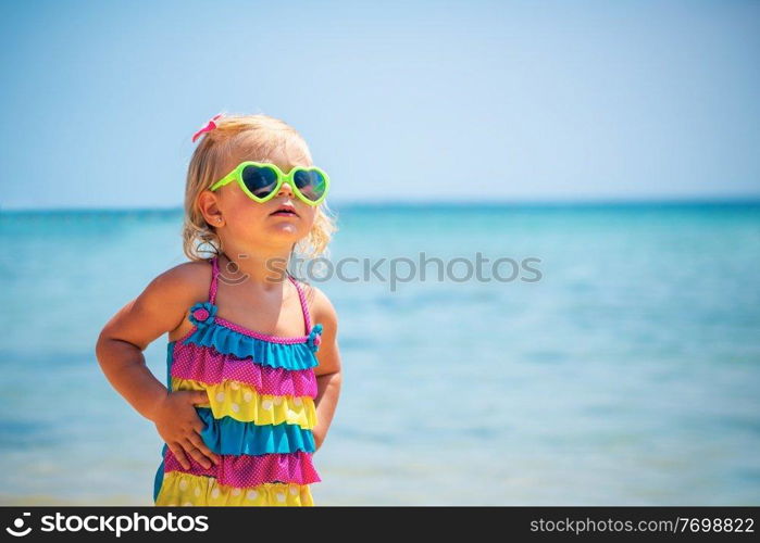 Cute little blond girl on the beach wearing sunglasses and stylish colorful swimsuit, child&rsquo;s fashion, summer vacation near the sea
