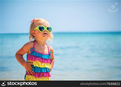 Cute little blond girl on the beach wearing sunglasses and stylish colorful swimsuit, child&rsquo;s fashion, summer vacation near the sea