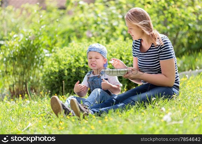 Cute little boy being fed outdoors on the grass by his mother reacting unfavorably to the food pulling a comical face