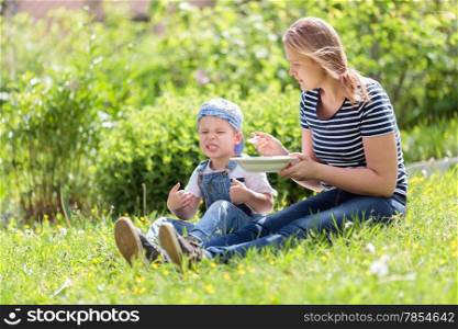 Cute little boy being fed outdoors on the grass by his mother reacting unfavorably to the food pulling a comical face