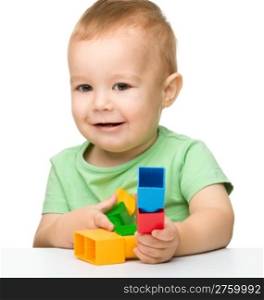 Cute little boy is playing with building bricks while sitting at the table, isolated over white