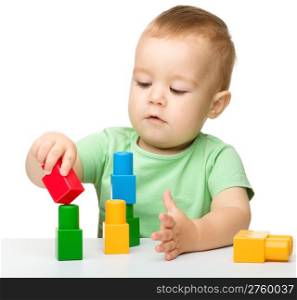Cute little boy is playing with building bricks while sitting at the table, isolated over white