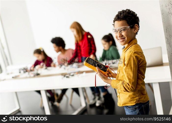 Cute little boy standing in front of  group of kids programming electric toys and robots at robotics classroom