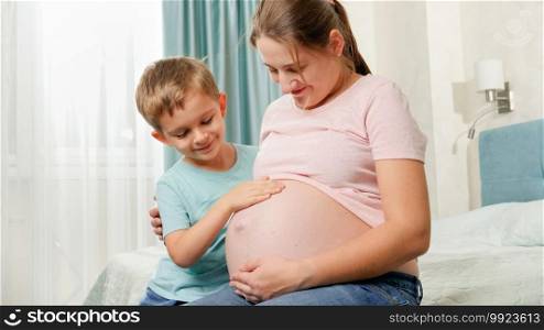 Cute little boy touching big belly of his pregnant mother sitting on bed at bedroom. Positive emotions in family expecting baby.. Cute little boy touching big belly of his pregnant mother sitting on bed at bedroom. Positive emotions in family expecting baby