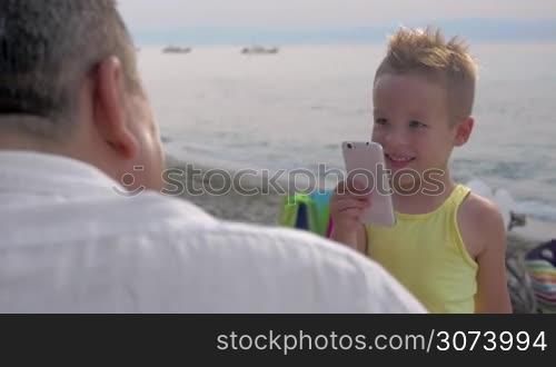Cute little boy with smart phone trying to make photo of grandparents on the beach. He is happy to take nice picture