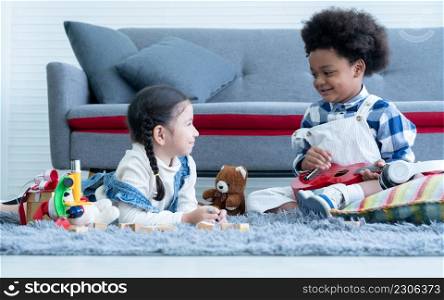 Cute little Caucasian and African kids girl and boy lying and sitting on floor smiling and playing toys build wooden blocks and ukulele together at home. Friendship of diverse ethnicity children