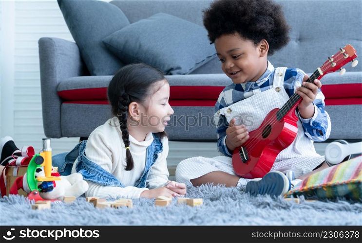 Cute little Caucasian and African kids girl and boy lying and sitting on floor singing and playing ukulele and toys together at home. Friendship of diverse ethnicity children