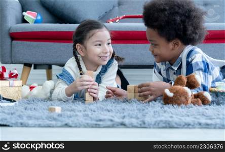 Cute little Caucasian and African kids girl and boy lying on floor smiling and playing toys build wooden blocks together at home. Friendship of diverse ethnicity children