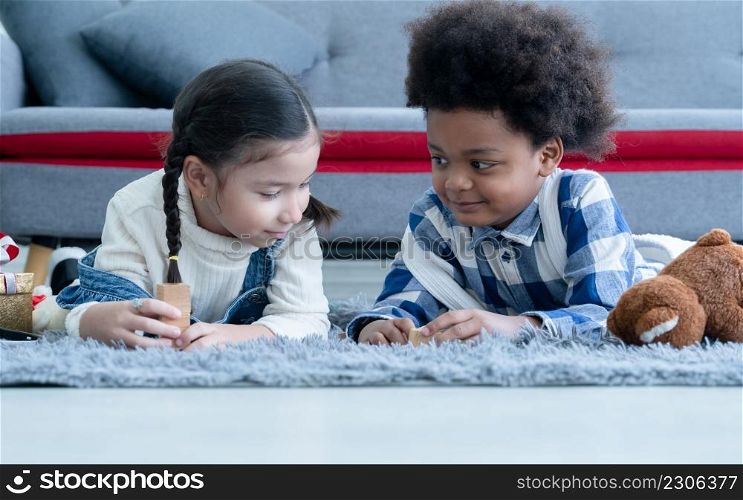 Cute little Caucasian and African kids girl and boy lying on floor smiling and playing toys build wooden blocks together at home. Friendship of diverse ethnicity children