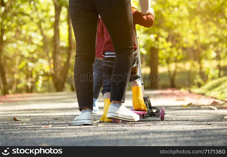 Cute little child girl having fun to riding a scooter on road with her mother in a summer park. Concept of happy family and childhood. Close up shot of legs of people in garden.