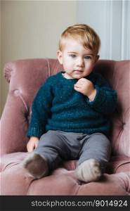 Cute little child with plump cheeks, blonde hair, wears fashionable clothes, sits on armchair, has concentrated look into camera. Curious kid with round face, poses, feels comfortably in chair