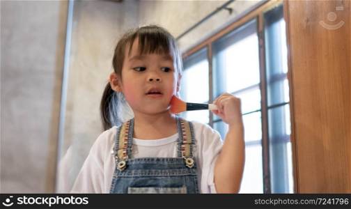 Cute little girl doing makeup and having fun brushing on cheek at a mirror in room.