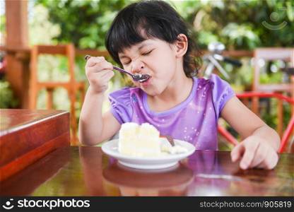 Cute little girl eating cake / Asian child happy and holding a spoon into the mouth with cake on dining table , selective focus