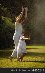 Cute little girl having fun with water under irrigation sprinkler with her mother