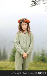 Cute little girl in a pastel jumpsuit with a colorful wreath on her head. Portrait of a girl in nature. A little girl outdoors in the foggy mountains looking in the camera. copy space.. Cute little girl in a pastel jumpsuit with a colorful wreath on her head. Portrait of a girl in nature. A little girl outdoors in the foggy mountains looking in the camera. copy space
