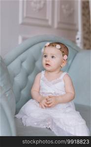 cute little girl in white dress and hoops of flowers on head is sitting on a couch in a classic style and having fun at home. cute little girl in white dress and hoops of flowers on head is sitting on a couch in a classic style and having fun at home.