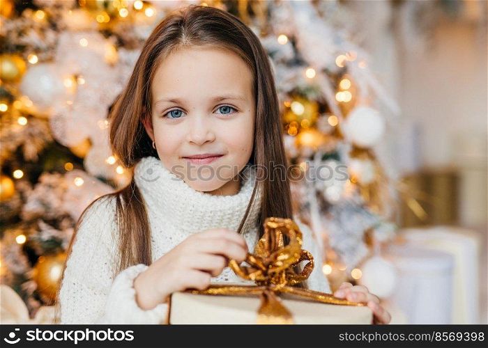 Cute little girl in white sweater sitting under Christmas tree and holding gift box.. Girl with her present